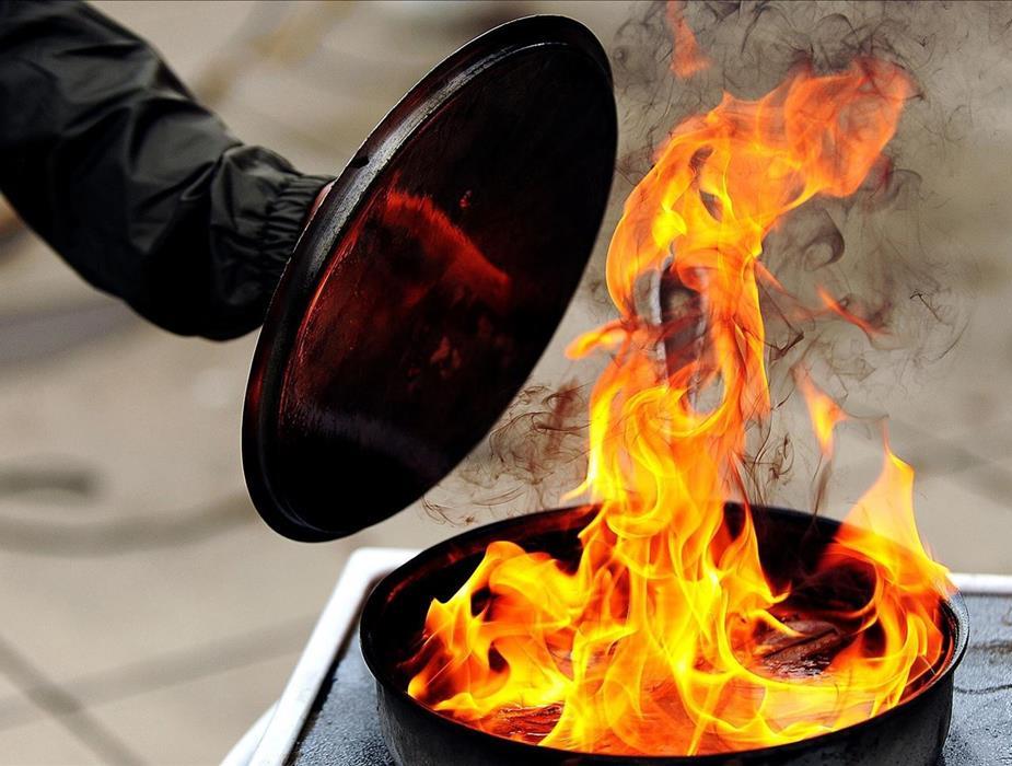 Grease fire in a pan, on the stove top. Flames in a pan with a person extending a metal lid to smother the fire.