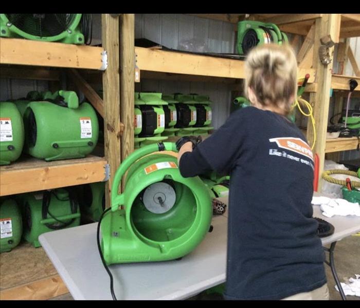 Several air movers on shelves inside building, employee with black SERVPRO shirt grabbing air mover off of grey table